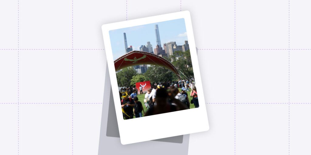 Outdoor event at a park with a crowd of people gathered under a red bandshell, with a Team Valor flag from Pokémon GO displayed prominently. The city skyline looms in the background under a clear blue sky, indicating an urban setting. The photo has a Polaroid frame, suggesting a snapshot of a community gaming event.